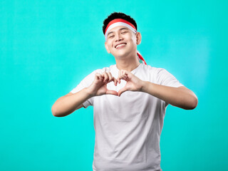 Naklejka premium A portrait of an Asian man celebrating Independence Day, wearing a white shirt with a red and white headband, making a heart-shaped pose in front of his chest, isolated with a turquoise background.