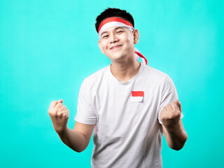 Naklejka premium A portrait of an Asian man celebrating Independence Day, wearing a white t-shirt and a red and white headband, showing a victory pose with his hands raised up. Isolated with a turquoise background.