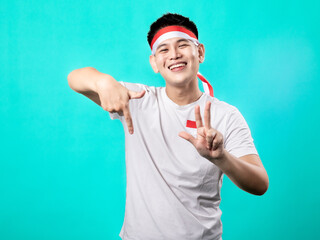 Naklejka premium A portrait of an Asian man celebrating Independence Day, wearing a white shirt with a red and white headband, posing with his fingers forming the number 78, isolated with a turquoise background.