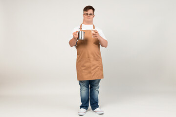 Smiling young man with down syndrome in glasses and barista uniform wearing apron and coffee in...