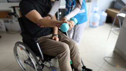 Paralyzed man sit in wheelchair and hold blue dumbbell on his outstretched arm. Femail doctor help patient to knead his hand in clinic.