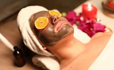 Woman undergo wellness course and face cleansing. Client lye on massage table with his hair wrapped in towel, coal mask on her face, and orange circles in front of her eyes.