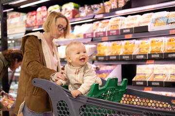 Caucasian mother shopping with her infant baby boy child choosing products in department of supermarket grocery store