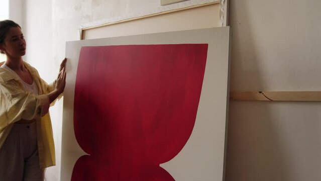 A young artist arranges her paintings in her studio. The woman carries a large canvas and places it against the wall. Workshop of a contemporary artist, abstract painting, art.