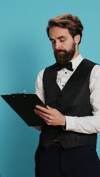 Vertical video Doorman verifying checklist of guests, holding register log on clipboard to check hotel visitors in studio. Employee checking reservations on official ledger for archive, taking notes.