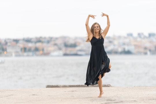 An adult smiling woman in black dress dancing ballet on the pier in cloudy weather