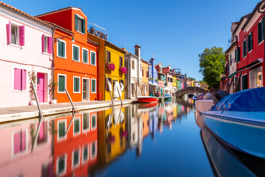 Colourful houses and buildings with reflection in blurred long exposure water of the river. Dock with moored boats on glassy water. Mirror of colors and bright vivid blue sky. Burano, Venice, Italy