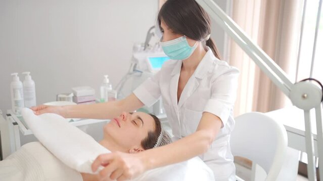 Beautician wraps female warm towel. Beauty concept. A cosmetologist performs cosmetic procedures to rejuvenate the face and body at a cosmetology clinic