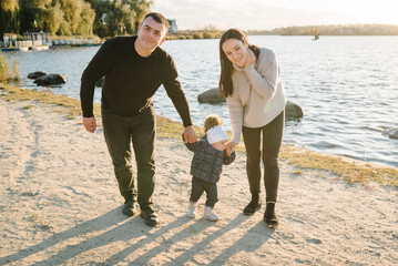 Family spending time together on beach at sunset on vacation. Mom and dad hold hands child walking in park. Concept of autumn holiday. Mother, father and daughter near lake in nature. Family photo.