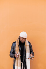 Fototapeta premium Happy stylish African American male in leather jacket hat and scarf browsing mobile phone while standing against brown wall