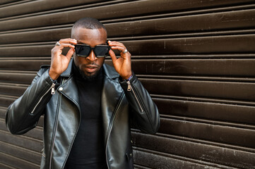 Serious masculine African American male in black leather jacket lowering stylish sunglasses and...