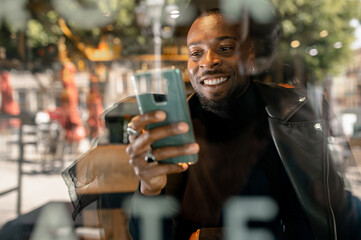 Stylish happy African American male sitting in cafe and surfing Internet on mobile phone