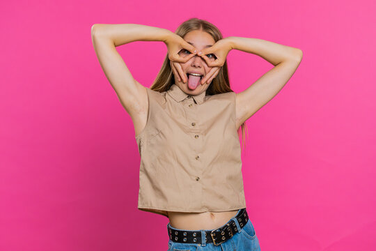 Funny comical playful young woman making silly facial expressions and grimacing, fooling around, showing tongue, idiotic expression. Girl isolated alone on pink studio background. People lifestyles