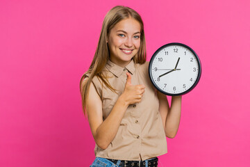 It is your time. Young attractive woman showing time on wall office clock, ok, thumb up, nice good, approve, start, pointing finger at camera. Blonde girl isolated on pink studio background, indoors