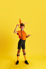 Full-length portrait of cheerful little boy, child in stylish clothes posing with smile against yellow studio background. Concept of childhood, kids emotions, fashion, hobby, ad