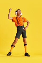 Fototapeta na wymiar Full-length of little boy, child in stylish clothes listening to music in headphones and singing against yellow studio background. Concept of childhood, kids emotions, fashion, hobby, ad
