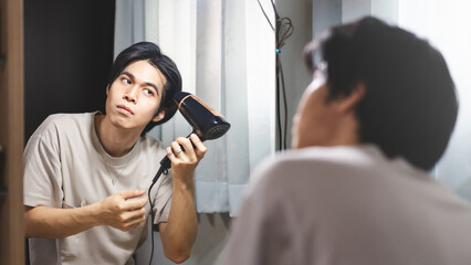 Young adult asian man using hair dryer blow for hairstyle in this mirror reflection at home