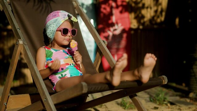Funny cute girl on summer vacation. The child has fun on the beach. Cute baby girl in a colorful swimsuit and sunglasses is resting and eating ice cream.