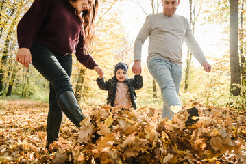 Mom and dad hold hands with child walking in yellow leaves in park. Family spending time together at sunset. Concept of autumn holiday. Mother father daughter run in forest. Bottom view down of legs.