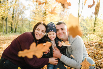 Fototapeta premium Autumn family photo. Happy family walks in autumn park. Mom, dad and daughter stand and hug on background of yellow leaves in forest. Upper half. Closeup. Young family spending time together at sunset