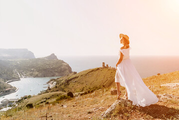 Fototapeta na wymiar Happy woman in a white dress and hat stands on a rocky cliff above the sea, with the beautiful silhouette of hills in thick fog in the background.