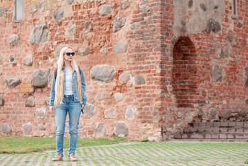 Obraz na płótnie Canvas Blonde girl in sunglasses with a backpack in jeans clothes on the background of a brick building.