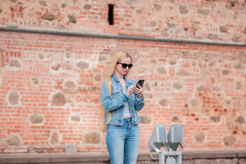 Blonde girl in sunglasses looking at the smartphone on the background of a brick building.