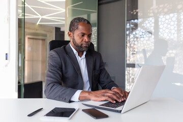 Serious mature businessman working on laptop inside office at workplace, african american boss thoughtful in business suit, successful experienced investor banker.