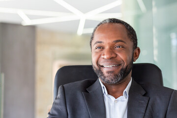 Close up of successful dreamy businessman smiling and looking at camera, african american boss in business suit smiling and looking at window inside office.