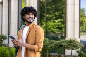 A young Indian man with a backpack and a phone in his hand stands on a city street and looks away with a smile. travels waiting to meet