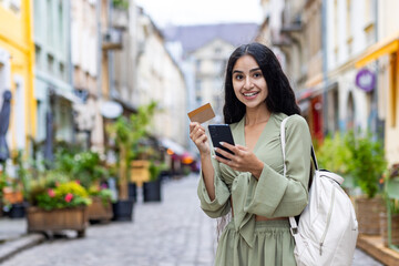 Beautiful young Indian woman using phone and credit card. Standing outside on a city street and smiling at the camera