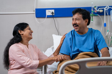 Happy smiling Indian wife talking with recovered husband near bed at hospital ward - concept of...