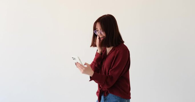 A Caucasian teenage girl on white backdrop. With a beaming smile, she using mobile phone. Suddenly, an unexpected discovery leaves her completely astounded. Her eyes widen, and she exclaims a Wow.