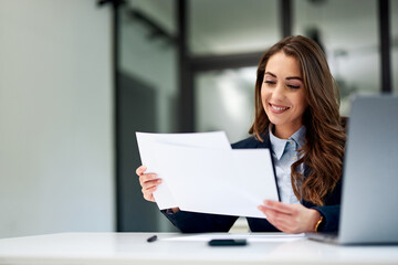 A smiling female employee is doing paperwork and using a laptop at the office.