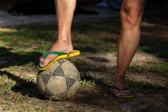 Mulher com um p&eacute; em cima de uma bola de futebol, cal&ccedil;ada com chinelos verde e amarelo em campo de v&aacute;rzea.