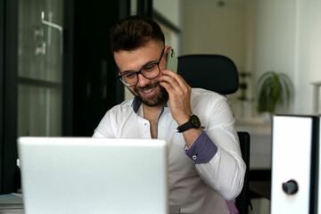 Portrait of smiling man with beard sitting with laptop, talking on phone
