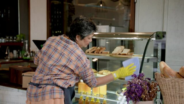 Elderly asia person who is mother bakery cafe owner, using glass cloth clean cake bakery cabinet, help daughter run cafe when she's busy, or maybe do something else about store.