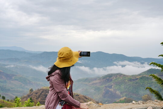 Tourists use their mobile phones to take pictures of fog-shrouded mountain views. Woman wearing purple-brown robe wearing yellow wide-brimmed hat and trousers with Red elephant pattern sit on chair.
