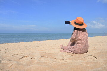 A female tourist wearing a brown skirt and orange hat sits on the sand and uses a mobile phone camera to capture the beauty of the sea and sky on a sunny day. The concept of relaxing on vacation.
