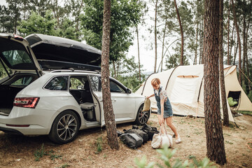 Woman packing things to her car roof storage at forest campsite after camping in tent. Travel and wilderness exploration experience. 