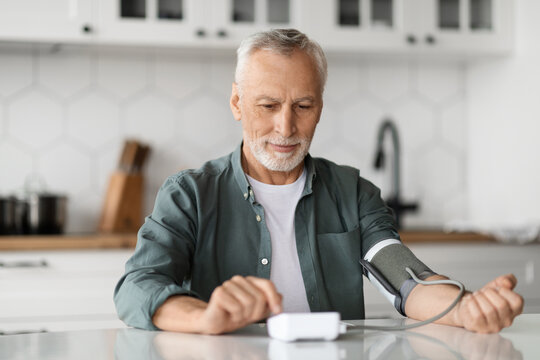 Senior Man Checking Blood Pressure With Upper Arm Monitor In Kitchen Interior