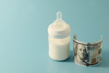 Baby bottle with formula or milk, next to $100. The concept of expensive artificial nutrition for babies. Blue background. Copyspace.