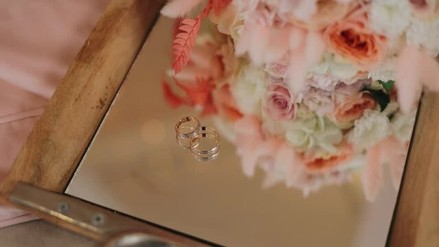 There is a bouquet of flowers and two wedding rings on a mirrored tray. The camera moves smoothly taking pictures of festive items