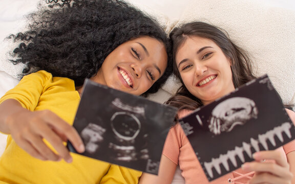 Portrait two diverse LGBT lesbian or female friends wearing casual clothes, laying together on bed in dark bedroom, showing baby ultrasound film, smiling with happiness. Pregnancy, Love Concept.