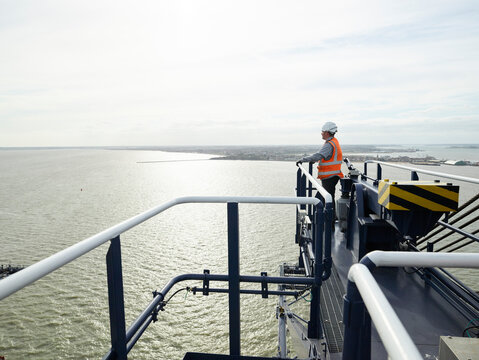 Dock worker looking at view from crane railing above cargo ship island view in background
