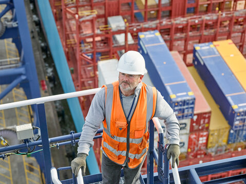 Looking down on dock worder supervisor going down railings with container ship and cargo in background