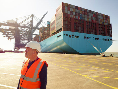 Dock worker wearing hard hat and reflective vest with cargo ship in background, Felixstowe, England.