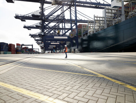 Dock worker standing beneath cargo crane and ship in shadows, Felixstowe, England.