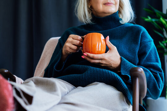 Cropped mature middle aged woman in sweater and warm socks relaxing in armchair with pumpkin shaped cup of hot coffee or tea drink. Cozy calm autumn holidays at home. Fall hygge mood concept