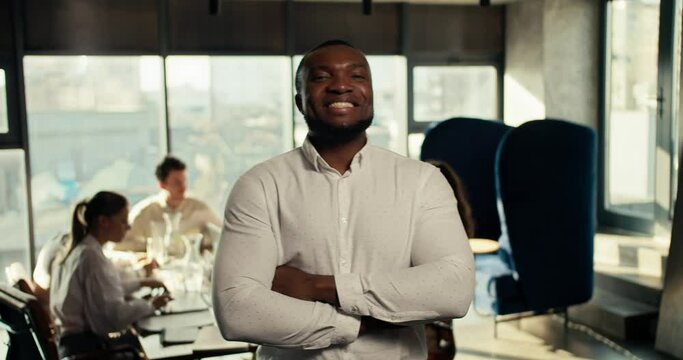 A successful businessman with Black skin in a white shirt folded his arms on his chest, smiles and looks at the camera against the backdrop of an office team at work